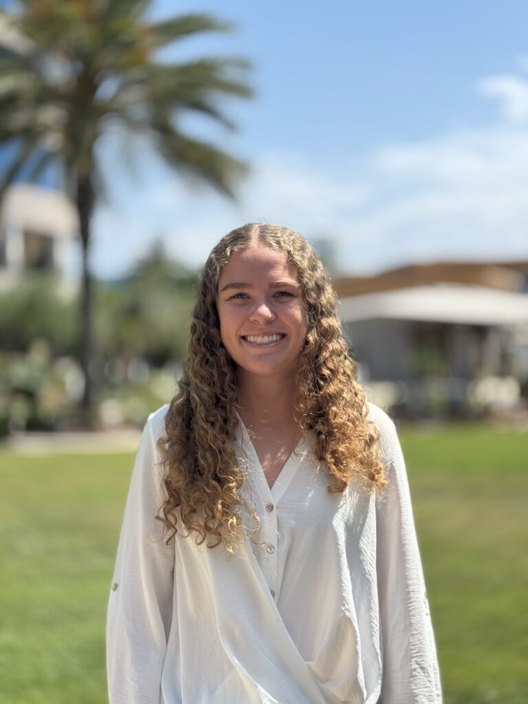 Headshot of Lucy Granger — Smiling woman with long curly hair in a white blouse with a blurred outdoor palm tree background.