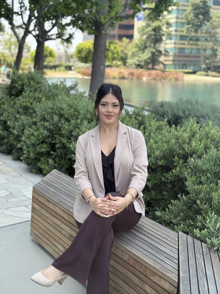 Isabella Reynoso sitting on a wooden bench in an outdoor park setting with a modern building and pond in the background.
